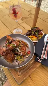 A short, static-heavy video showcasing a meal of hot honey chicken and beef loaded fries, accompanied by a Lillet spritz, at an outdoor venue.