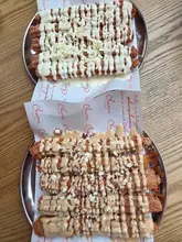 A top-down, overhead shot of two metal plates serving churros. One plate features churros drizzled with white chocolate sauce and white chocolate shavings, while the other features churros with a light brown sauce and crushed chocolate pieces, served on branded paper liners.
