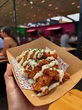 A close-up, first-person perspective shot of a cardboard tray filled with crispy fried chicken strips drizzled with a creamy garlic herb sauce, held in a hand at an outdoor street food venue.