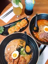 A high-quality, top-down overhead shot of a Japanese dining spread featuring two bowls of ramen, a plate of dynamite prawns, and two vibrant cocktails on a wooden table.
