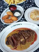 A high-quality, top-down food photography shot featuring a steak with peppercorn sauce, a side of fries, and a macaroni pie served on a terrazzo-style table at Anderson's in Glasgow.