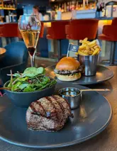 A table spread at a restaurant featuring a steak, a burger, a side salad, and a cocktail, showcasing a meal deal offer.