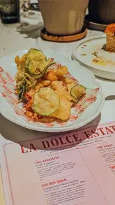 A close-up, slightly high-angle shot of a plate of fritto misto served on branded paper, positioned next to a printed menu at a restaurant table. The image captures the texture of the fried food, though the overall atmosphere is heavily influenced by warm, ambient indoor lighting.