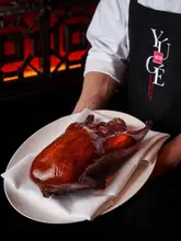 A professional, high-quality promotional shot featuring a chef presenting a whole roasted duck at Yú Gé Cantonese Cuisine. The lighting and composition highlight the texture and presentation of the dish, conveying a premium dining experience.