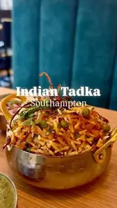 A close-up, eye-level shot of a vegetable biryani served in a traditional metal bowl, set against a blurred blue restaurant interior. The focus is sharp on the rice and garnish, presenting the dish in a clear and appealing manner.