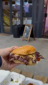 A casual, first-person perspective shot of a smash burger being held up, likely at a street food venue. The focus is on the burger, showcasing its messy, juicy texture, with a blurred background suggesting an outdoor dining setting.