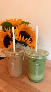 A casual, user-generated style photo of two iced drinks in takeaway cups from Coffee#1, placed on a wooden surface in front of a vase of sunflowers.