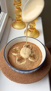A close-up, overhead shot of a bowl of baked oats topped with banana slices, with a salted caramel peanut butter sauce being poured over the top. The lighting is soft and natural, highlighting the texture of the food.