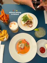 A top-down, flat-lay shot of an outdoor dining table featuring a crab gazpacho, grilled octopus, sweet potato fries, and cocktails. The image captures an alfresco dining experience in a garden setting.