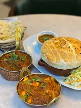 A top-down, slightly elevated view of a table spread at an Indian restaurant, featuring a large pastry-covered dish, curry bowls, and naan bread.