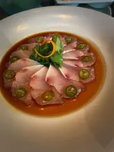 A high-angle, close-up shot of the signature Yellowtail Jalapeño dish at Nobu. The raw fish is arranged in a precise circular pattern on a large white plate, garnished with fresh cilantro and jalapeño slices, resting in a soy-based sauce.
