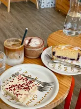 A casual, overhead shot of a coffee shop table featuring an iced latte, a hot chocolate, and two slices of cake on branded plates. The setting is simple and relaxed, typical of a local cafe visit.