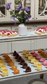A high-quality, aesthetically pleasing shot of an afternoon tea service at the Corinthia London, featuring a tiered stand filled with delicate pastries and a vase of purple flowers in the background.