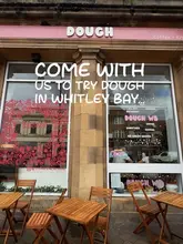 A casual exterior shot of a cafe called 'Dough' in Whitley Bay, featuring outdoor seating and a pink-themed storefront. The image is used to promote the venue as a spot for brunch, lunch, or coffee.