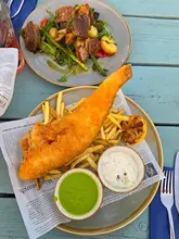 A bright, overhead shot of a pub-style dinner featuring a large portion of battered fish and chips alongside a lamb dish, served on a blue wooden table.