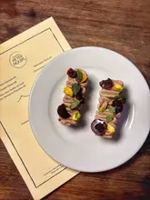 A top-down, overhead shot of two pieces of chicken liver parfait on toast, garnished with cherries and pistachios, served on a white plate alongside a menu from the restaurant.