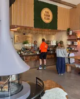 A wide-angle shot of the interior of 'The Farm Club', showing a wooden counter with a menu board, a fireplace in the foreground, and customers interacting with staff. The space has a rustic, modern aesthetic with wood paneling and greenery.