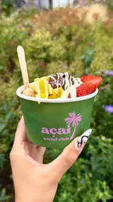 A bright, close-up shot of an açai bowl held in a hand, featuring a colourful arrangement of banana, strawberry, mango, Nutella, and white chocolate drizzle, set against a soft-focus outdoor background.