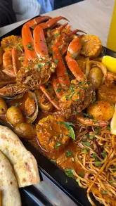 A close-up, top-down shot of a seafood boil platter featuring crab legs, corn, potatoes, mussels, shrimp, and noodles. The image captures the messy, communal nature of the dish, though the framing is slightly tight.