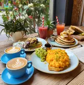 A bright, well-composed photograph of a brunch spread at a cafe, featuring scrambled eggs on toast, avocado toast, pancakes, and flat white coffees. The natural lighting and clean table setting create an inviting atmosphere.
