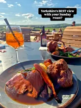 A high-quality, well-composed shot of a Sunday roast dinner served outdoors at a pub with a scenic countryside view. The focus is sharp on the roast beef and Yorkshire pudding, with a cocktail in the foreground and a blurred background of the beer garden, effectively capturing the atmosphere of a pleasant weekend meal.