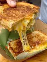 A high-quality, close-up shot of a grilled cheese sandwich being pulled apart, showcasing a long, appetizing cheese stretch. The sandwich is served in a branded paper tray on a wooden surface, with the background softly blurred to keep focus on the food.