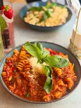 A close-up, top-down shot of a bowl of fusilli arrabbiata, garnished with fresh basil and a generous dusting of parmesan cheese. In the background, a second dish and a drink are visible, providing context for a dining setting.
