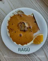 A high-quality, top-down food photography shot of a single scallop served in a shell with a rich bisque, accompanied by a slice of orange. The image is clean, well-lit, and focuses on the texture and presentation of the dish.