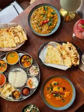 A high-quality, overhead flat-lay shot of an Indian feast featuring various curries, a thali platter, naan bread, and a cocktail, set on a warm wooden table.
