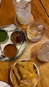 A top-down, overhead shot of a Mexican meal featuring a bowl of totopos, a metal tray with three different salsas, and a cocktail on a wooden table at a restaurant in Newcastle.