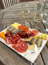 A top-down, natural light shot of a charcuterie board featuring various cured meats, cheeses, crackers, and a jar of chutney, served alongside two glasses of white wine on a rustic wooden table at a winery.
