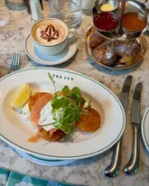 A high-quality, well-composed overhead shot of a breakfast spread at The Ivy, featuring a smoked salmon and potato rösti dish, a latte with latte art, and a side of pastries. The lighting is soft and natural, highlighting the textures of the food, while the marble table background provides a clean, upscale aesthetic.