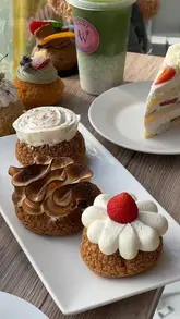 A visually appealing, well-lit shot of a variety of choux buns and a slice of cake, showcasing the patisserie's offerings. The natural lighting and shallow depth of field draw attention to the textures and details of the pastries.