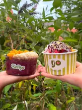 A bright, outdoor shot of two hands holding a takeaway acai bowl and a frozen yogurt cup against a background of green foliage.