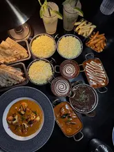 An overhead flat-lay shot of a generous Indian feast featuring multiple curries, rice bowls, naan bread, fries, and cocktails on a dark table.