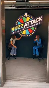 A promotional shot for 'BuzzAttack,' a game show experience in London, featuring two people sitting on stage chairs in front of the venue's branding.
