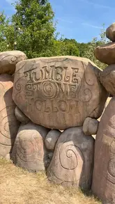 A bright, clear, and well-composed photograph of the 'Tumble Stone Hollow' sign at Stonor Park, effectively establishing the location for a family-friendly outdoor activity venue.