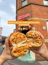 A high-quality, first-person perspective shot of two halves of a loaded sandwich held up in front of the iconic Afflecks building in Manchester, effectively showcasing the food and the location.
