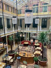 A high-angle, symmetrical architectural shot of a hotel atrium, featuring a glass roof, hanging pendant lights, and a central seating area with plants and tables.