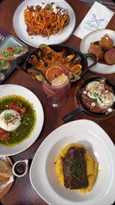 An overhead flat-lay shot of a table filled with a variety of dishes, including paella, pasta, burrata, croquettes, and sangria, showcasing a 'feast' style dining experience.