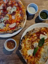 An overhead shot of two pizzas and several small dipping bowls on a wooden table, likely taken at a casual Italian restaurant. The image focuses on the food, showcasing the textures of the pizza toppings and the accompanying dips.