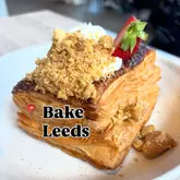 A close-up, high-quality shot of a layered pastry topped with cream, crumble, and a fresh strawberry slice, served on a simple plate. The image effectively showcases the texture and freshness of the product.