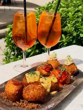 A bright, high-quality shot of a bottomless brunch spread featuring Aperol Spritzes and a variety of croquetas on a rustic wooden board, set against an outdoor backdrop.