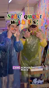 A high-energy, vlog-style video featuring two creators visiting a 'Paint Splat' venue in Manchester. They demonstrate various methods of painting, including using fly swatters, syringes, water guns, and a spin art turntable, while wearing protective ponchos.