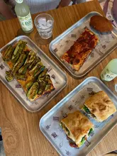 A bright, overhead flat-lay shot of a variety of Sicilian food items served on metal trays, including pizza, a sandwich, an arancino, and lemonade, set against a wooden table background.