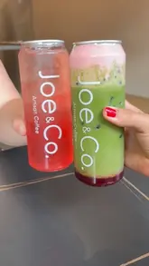 A simple, casual shot of two hands holding branded iced drinks from a cafe, featuring a strawberry iced tea and a strawberry matcha latte. The composition is straightforward, focusing on the product branding, though the framing is slightly tight.