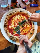A top-down, POV shot of a Margherita pizza being cut, set on a wooden table with children's toys in the background, effectively communicating a family-friendly dining experience.