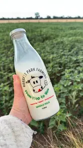 A clean, effective product shot featuring a glass bottle of whole milk from Rosset Park Farming, held against a soft-focus farm field background. The image successfully highlights the farm-to-table nature of the product with natural lighting and a simple, uncluttered composition.