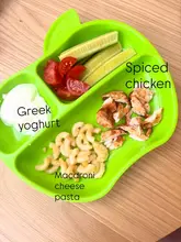 A clear, top-down, well-lit photograph of a toddler's meal served on a green, apple-shaped plastic plate. The image features distinct portions of cucumber, cherry tomatoes, Greek yoghurt, macaroni cheese, and spiced chicken, with text overlays identifying each component.