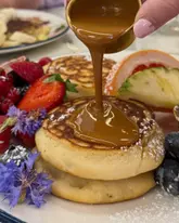 A high-quality, close-up action shot of sauce being drizzled over a stack of pancakes, garnished with fresh fruit. The image captures a classic brunch aesthetic with bright, natural lighting and a shallow depth of field that keeps the focus on the food.